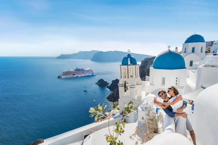 Ein Paar macht ein Selfie vor den blauen Kuppeln Santorins, während im Hintergrund ein großes Kreuzfahrtschiff in der Ägäis ankert.